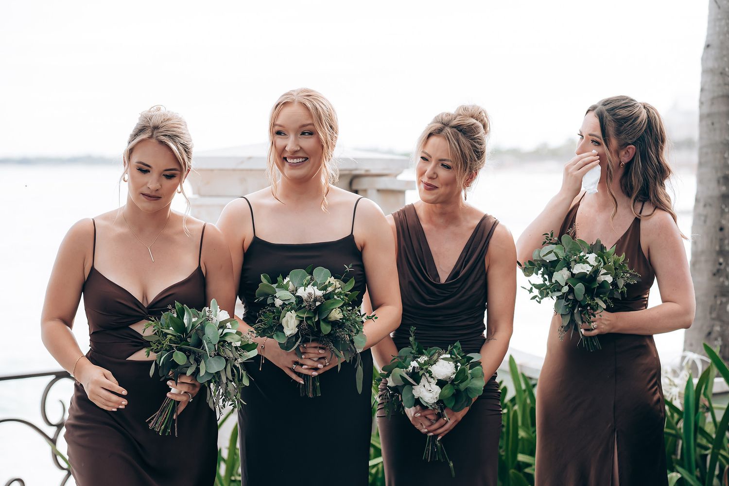 A group in dark green and black dresses hold bouquets while smiling and laughing together at an event.