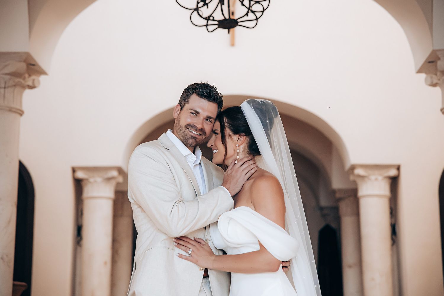 Romantic moment captured under elegant arched columns and chandelier in white interior.