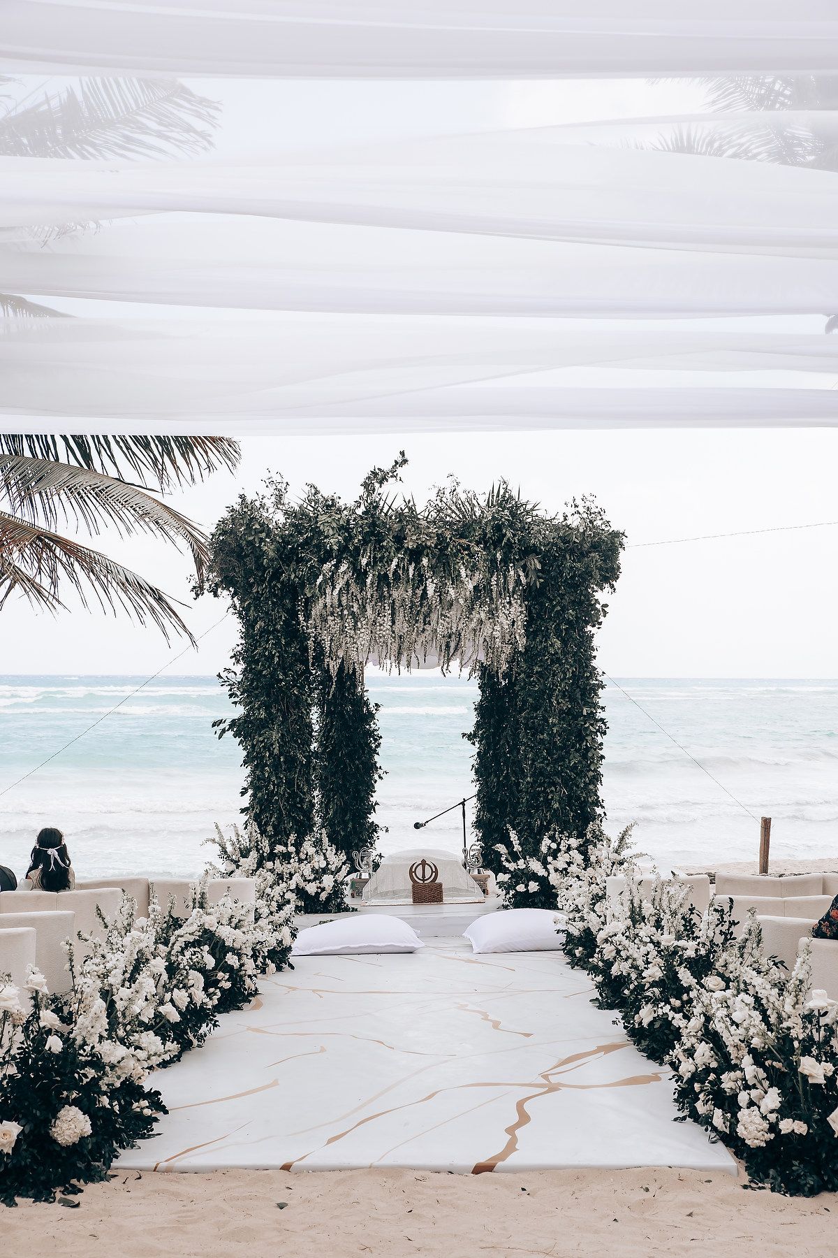 Elegant beach wedding ceremony setup with greenery arch and white floral arrangements.