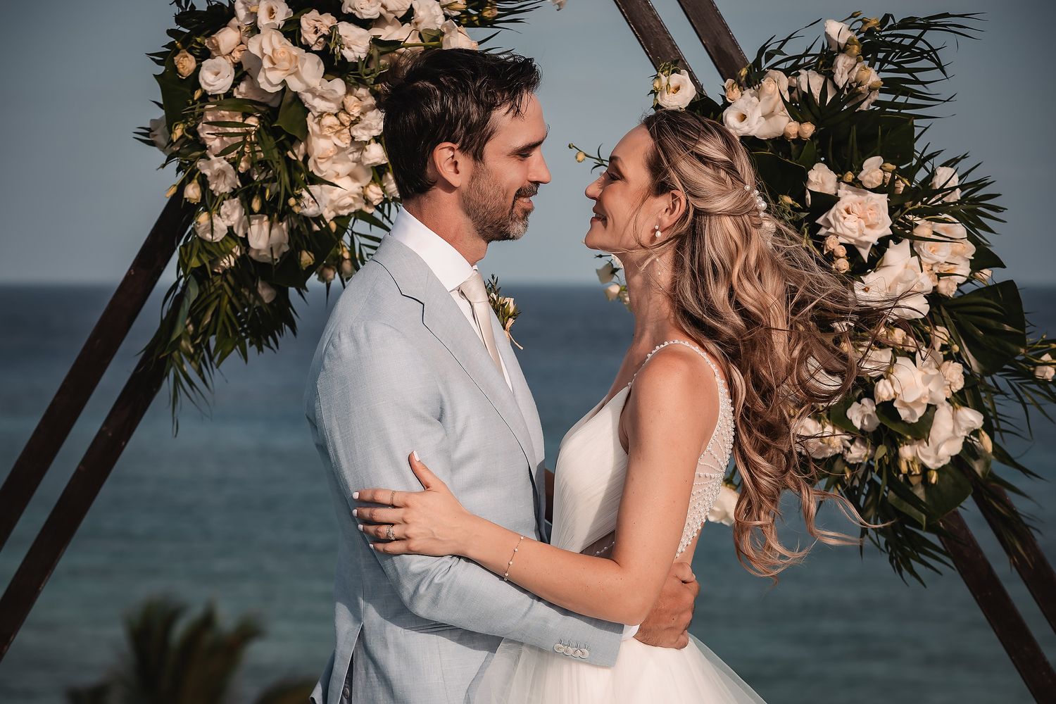 Wedding ceremony under a floral arch with ocean backdrop and romantic lighting.