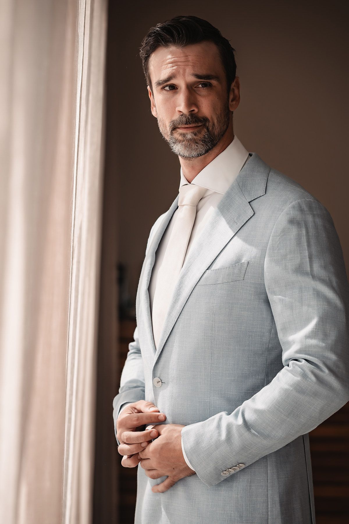 Person in light gray suit jacket posing against window light.