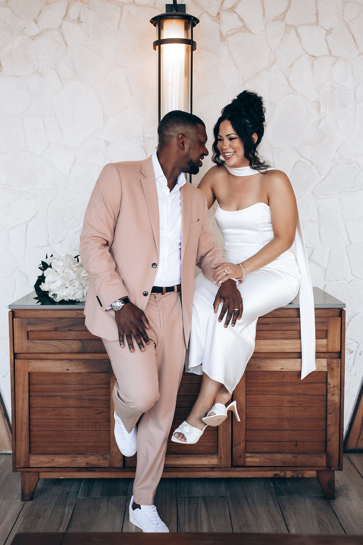 A couple shares a playful moment while sitting on wooden furniture under a wall lamp.