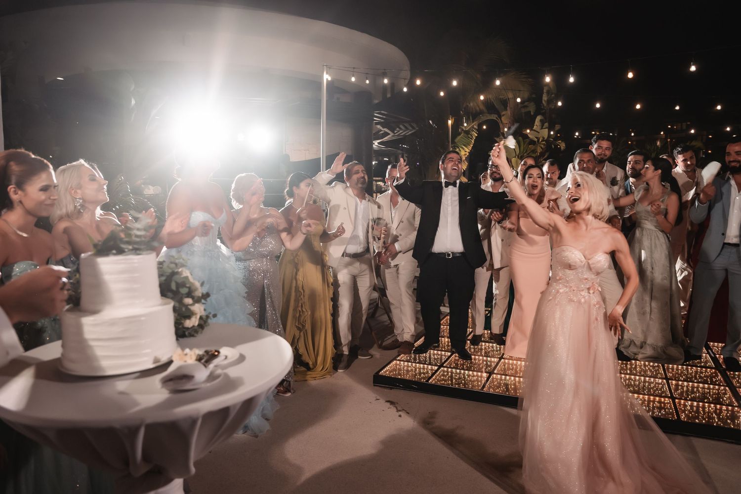 Wedding reception guests dancing and celebrating on a dimly lit dance floor under string lights.