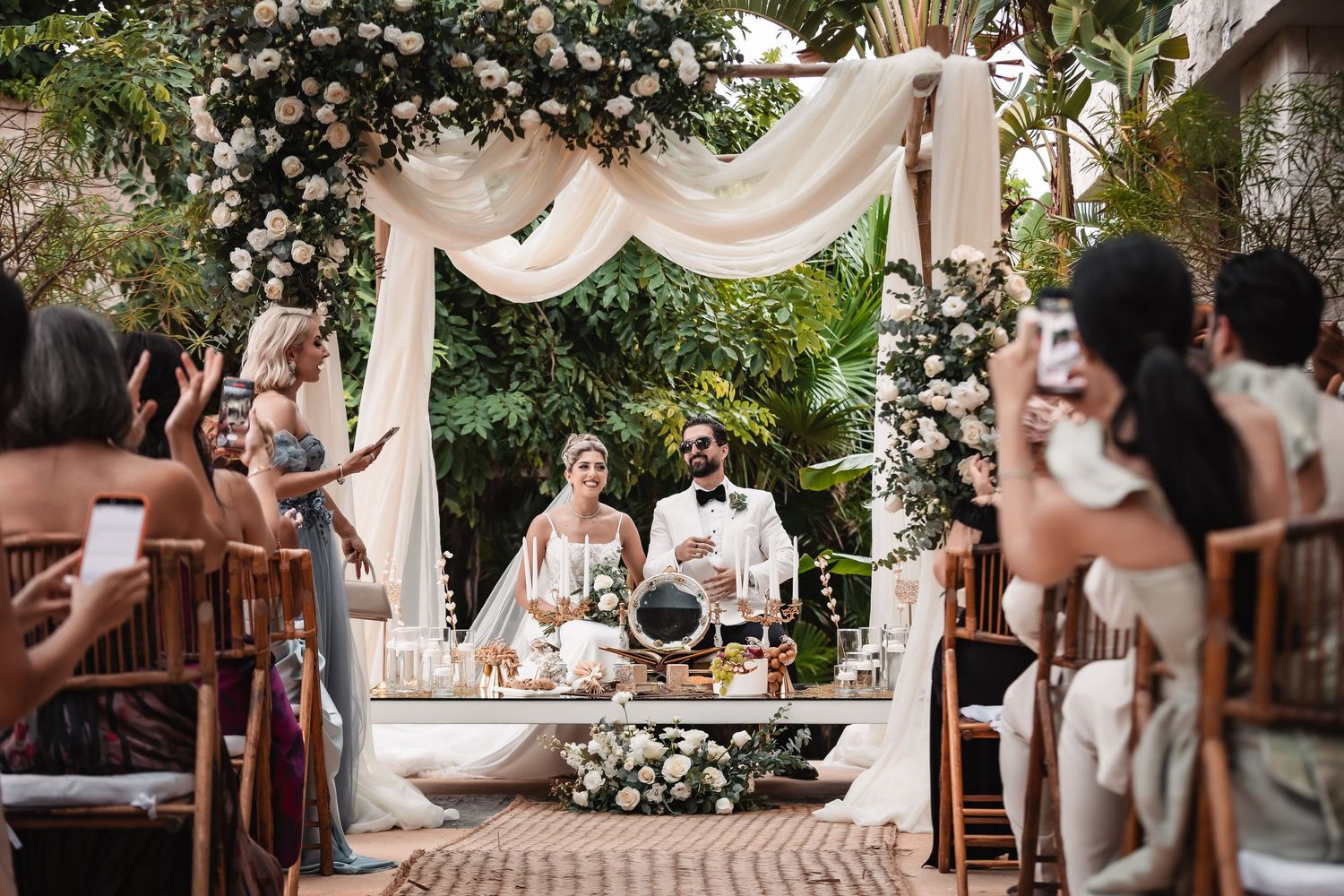 Outdoor wedding venue decorated with white drapes and floral arrangements in a garden setting.