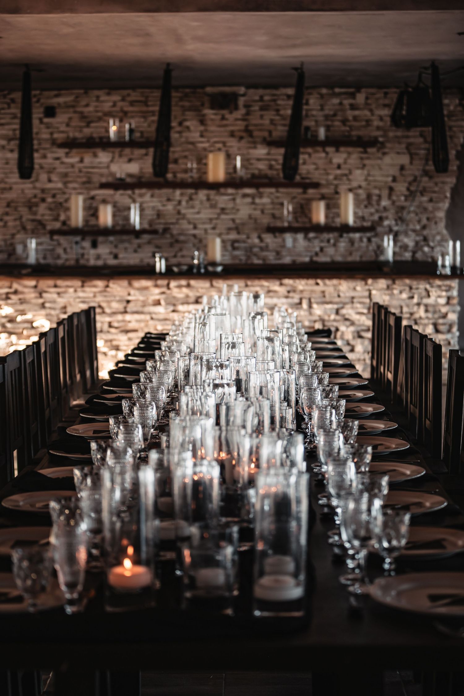 Dramatic table setting with multiple glass vessels and candles against rustic stone wall.
