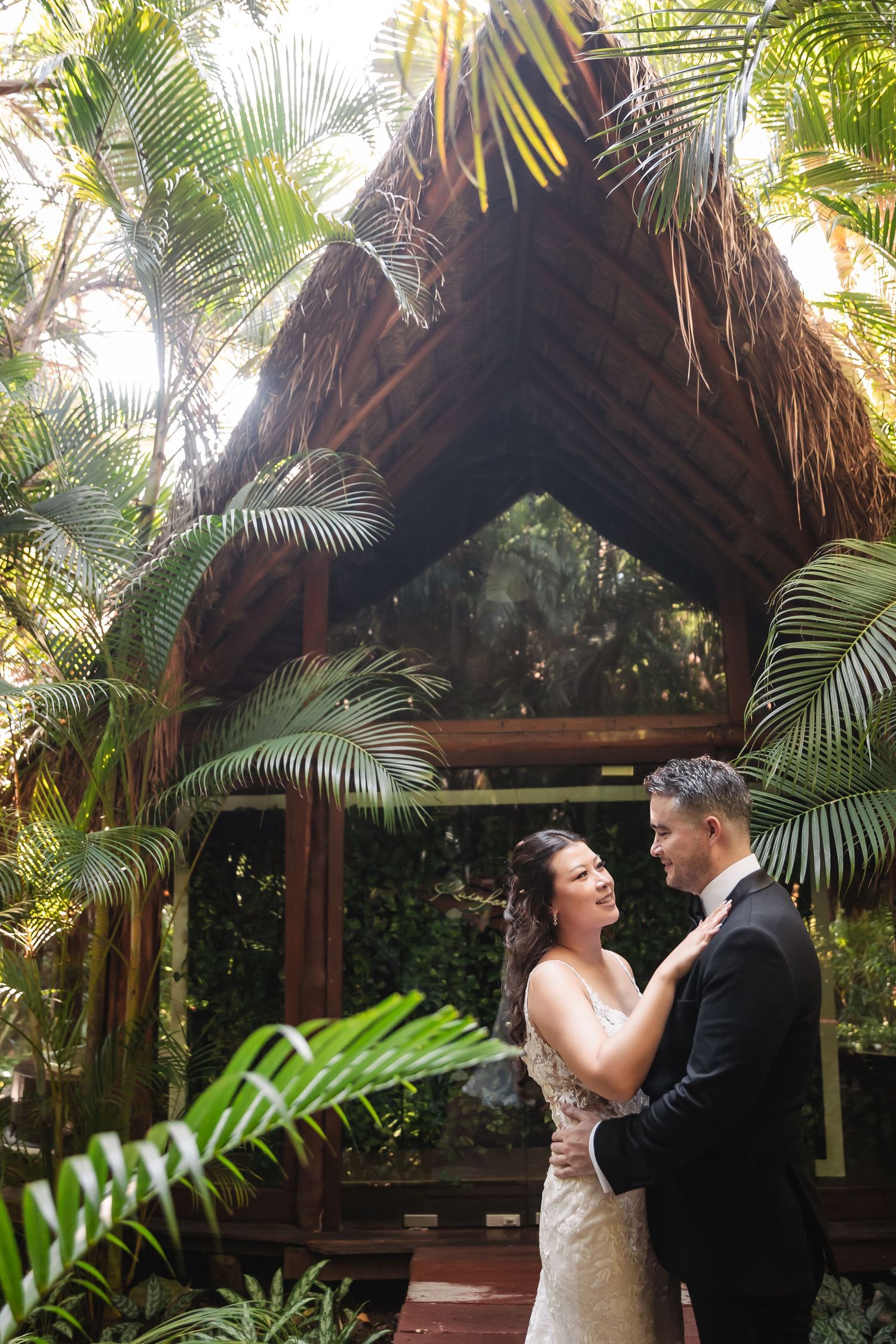 Wedding couple embraces under rustic thatched-roof gazebo surrounded by lush tropical palms.