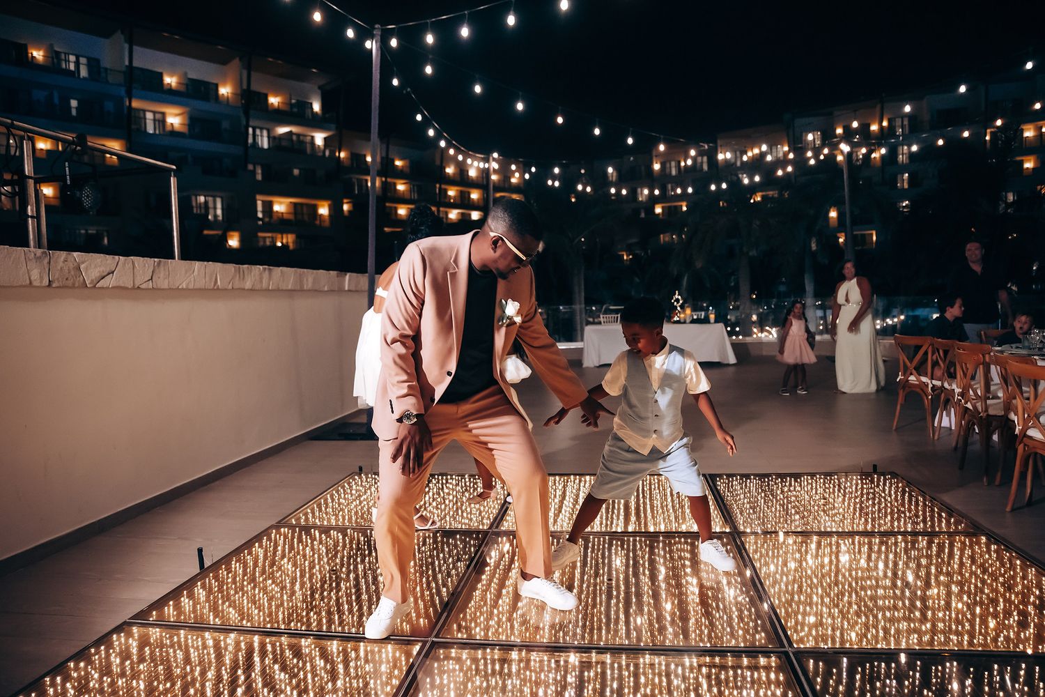 People dancing and celebrating on wooden platform under twinkling outdoor lights.
