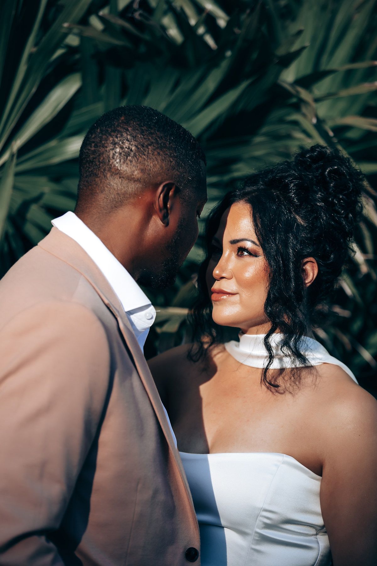 Romantic wedding couple in tan suit and white dress share intimate moments in a tropical garden setting.