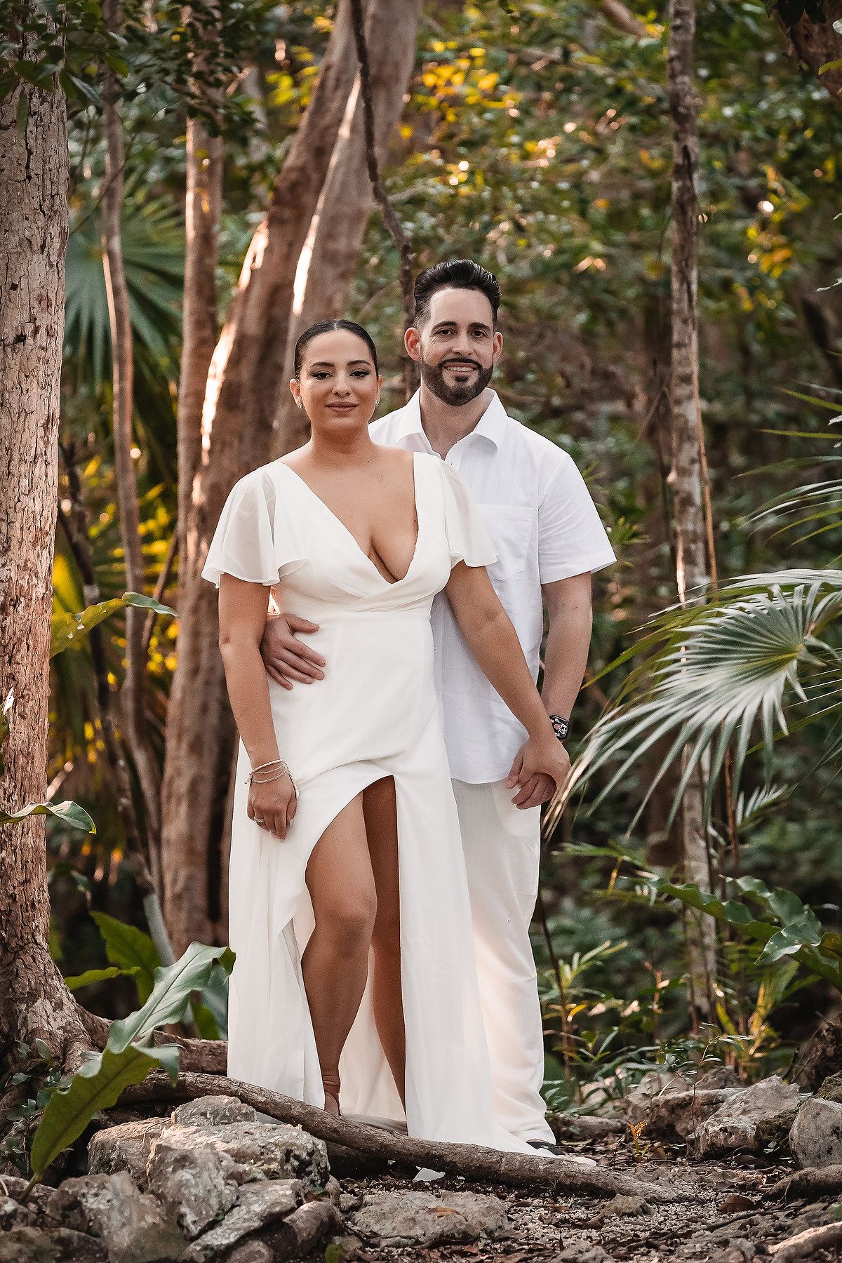 A romantic couple in white outfits poses together on rocky terrain surrounded by lush tropical foliage.
