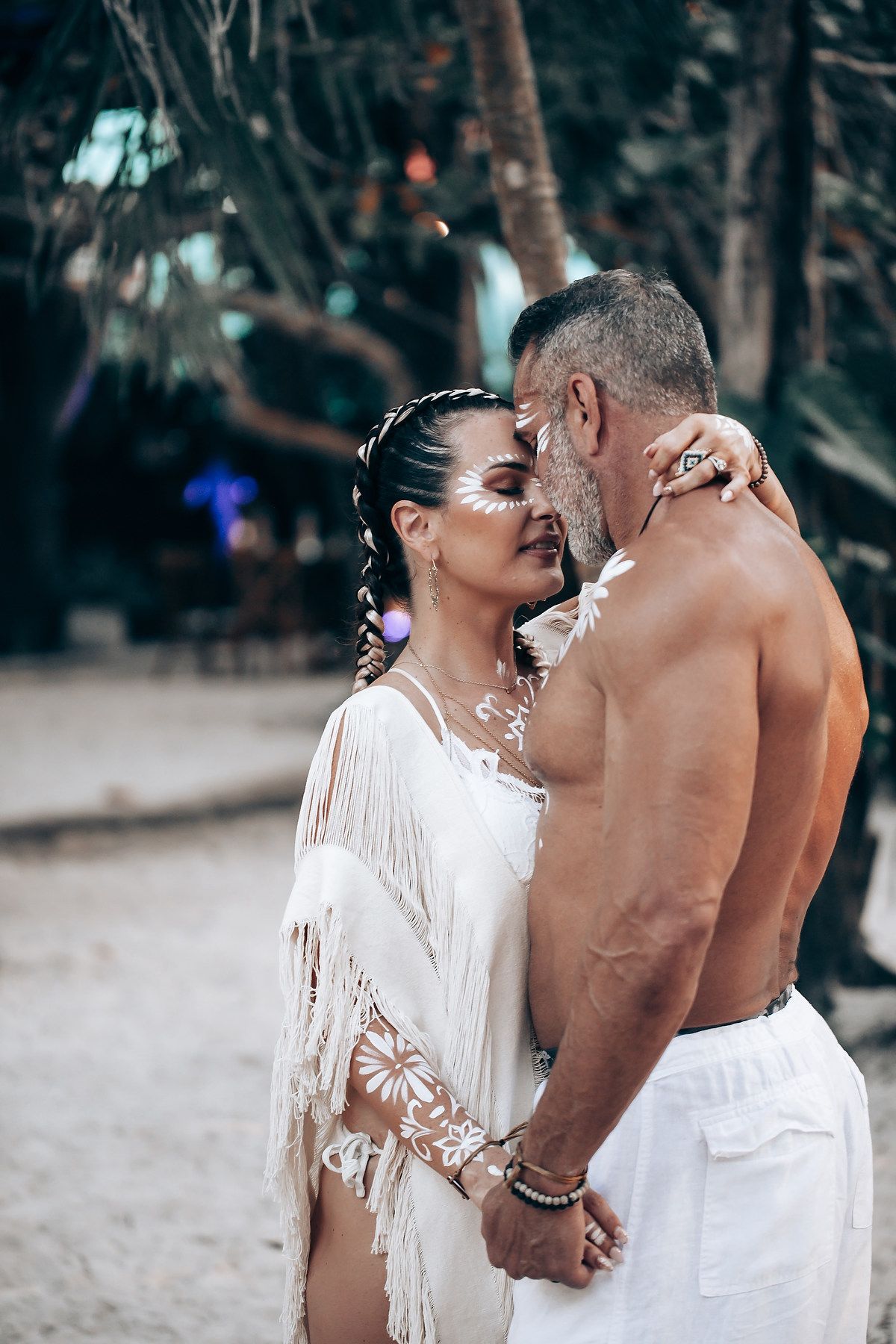 A romantic beach wedding photoshoot with white attire and body paint decorations in a tropical setting.