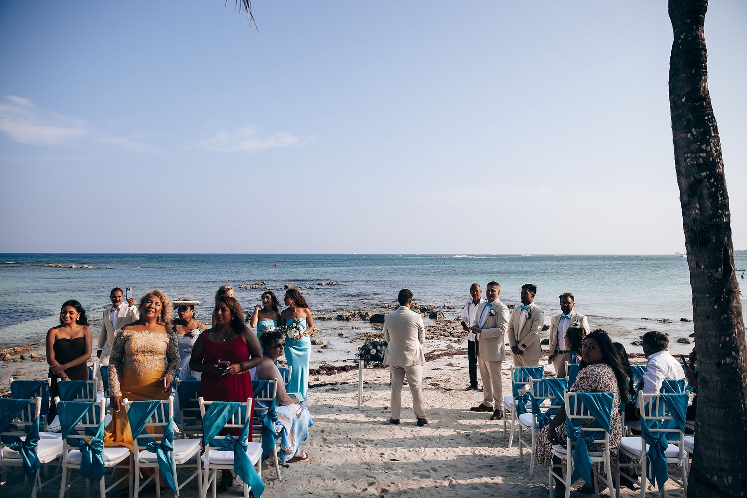 Guests seated at a beachfront wedding ceremony with palm trees and turquoise ocean views in the background.