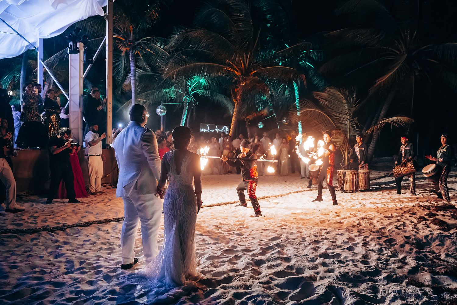 Beachside wedding celebration at night with ambient lighting and guests dancing on sandy shores.