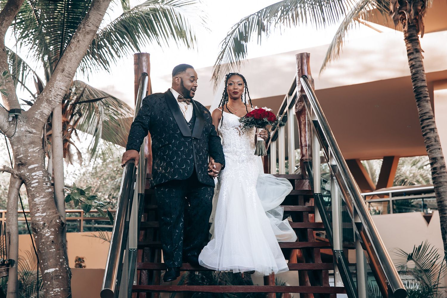 A newlywed couple descends an outdoor staircase surrounded by palm trees at a tropical venue.