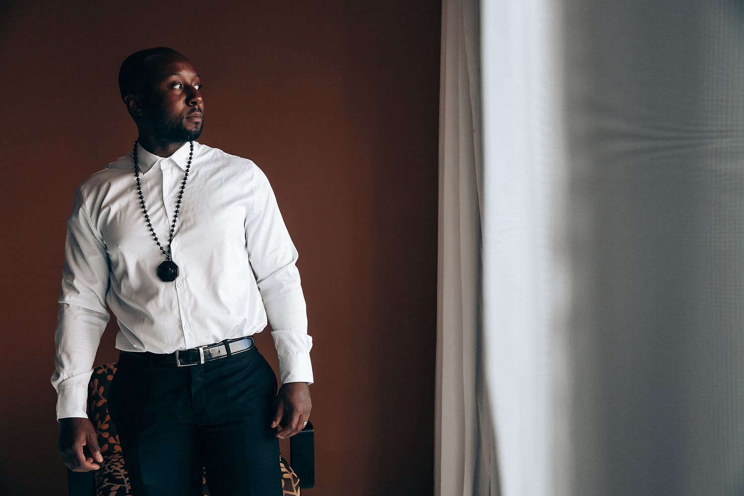 A person in a white dress shirt and dark pants stands confidently near a window.