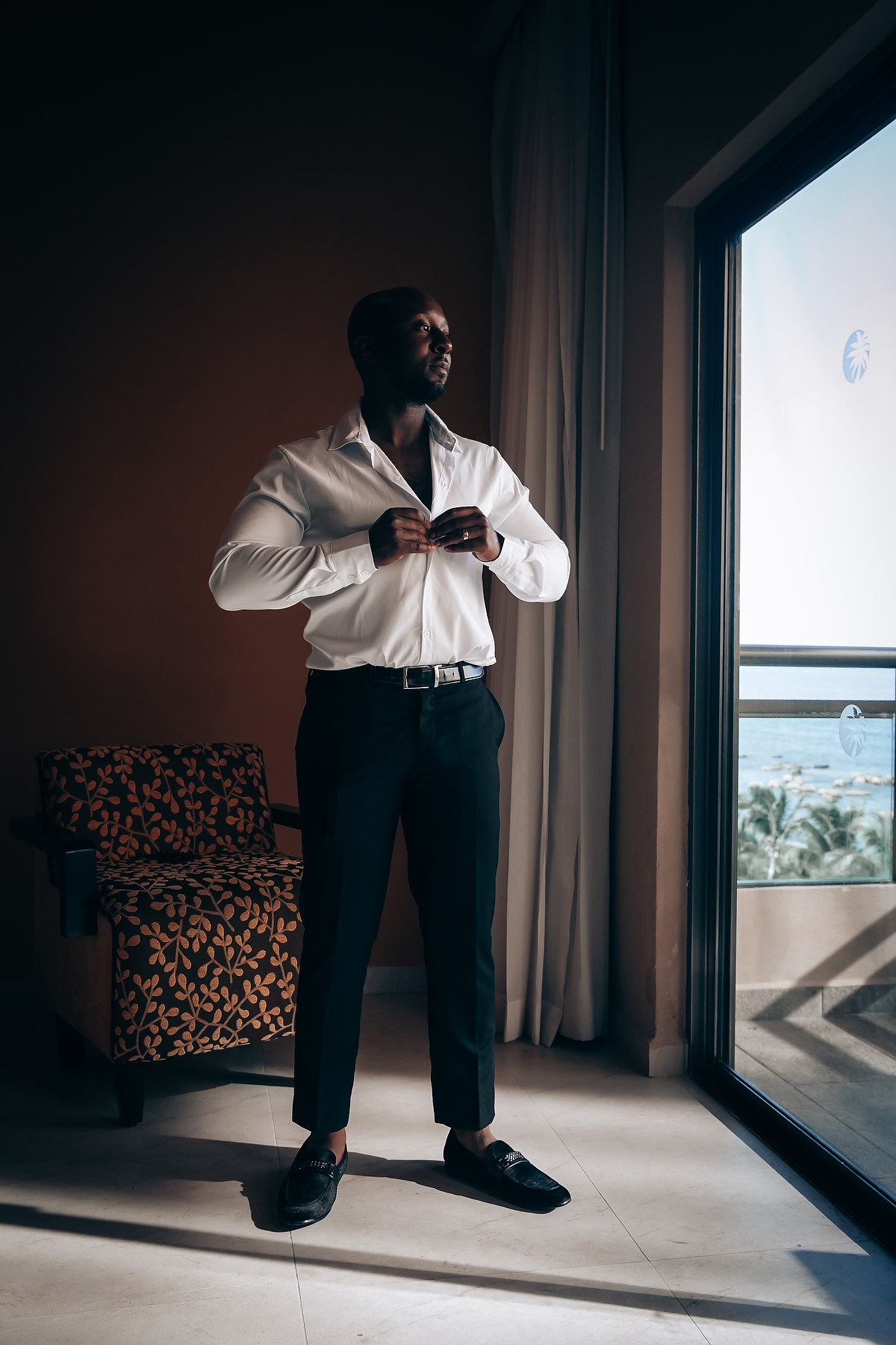 A person wearing formal attire adjusts their shirt while standing by a window with ocean views.