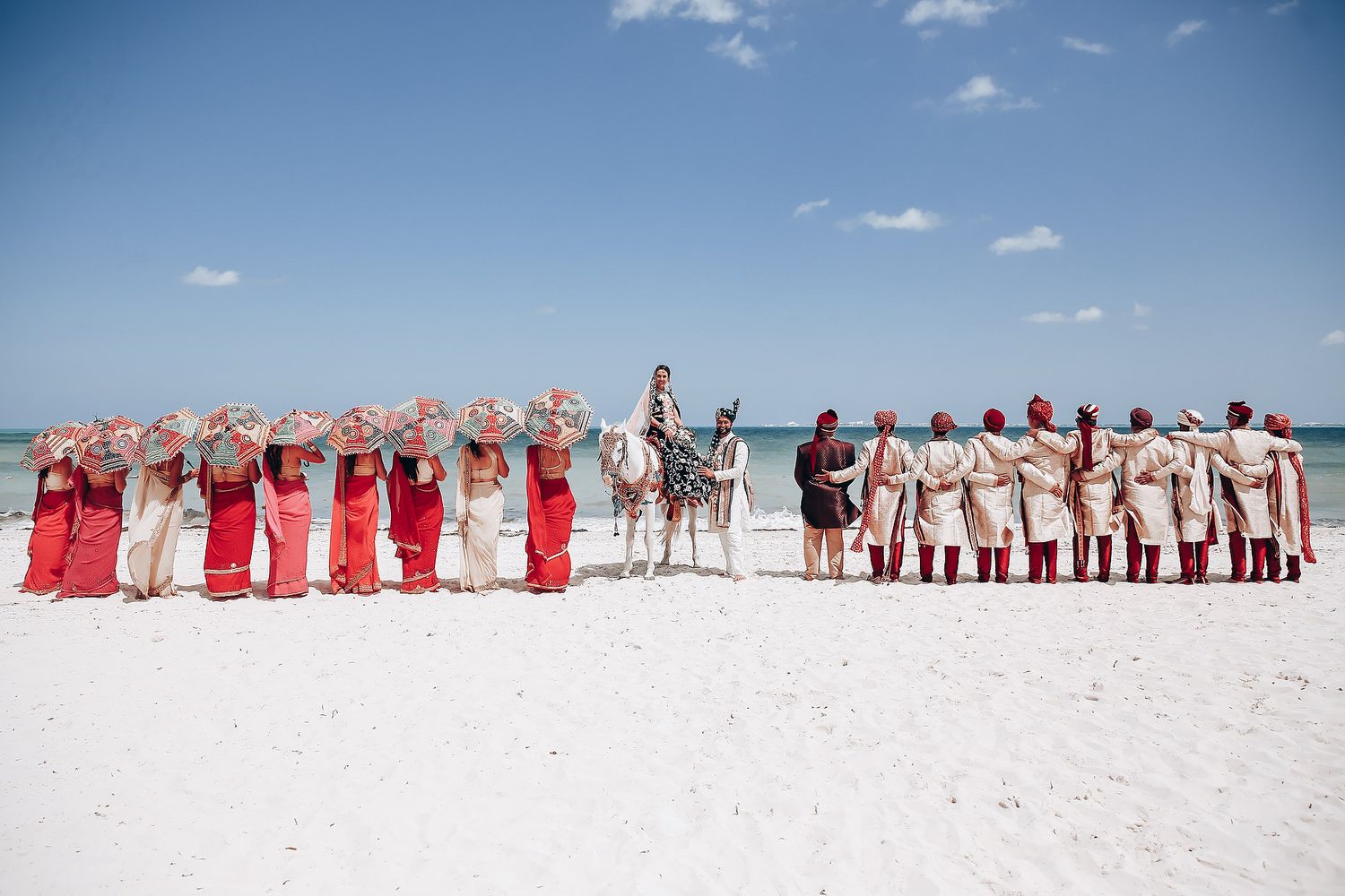 Wedding party members lined up on a white sandy beach wearing red and white attire against a blue sky.