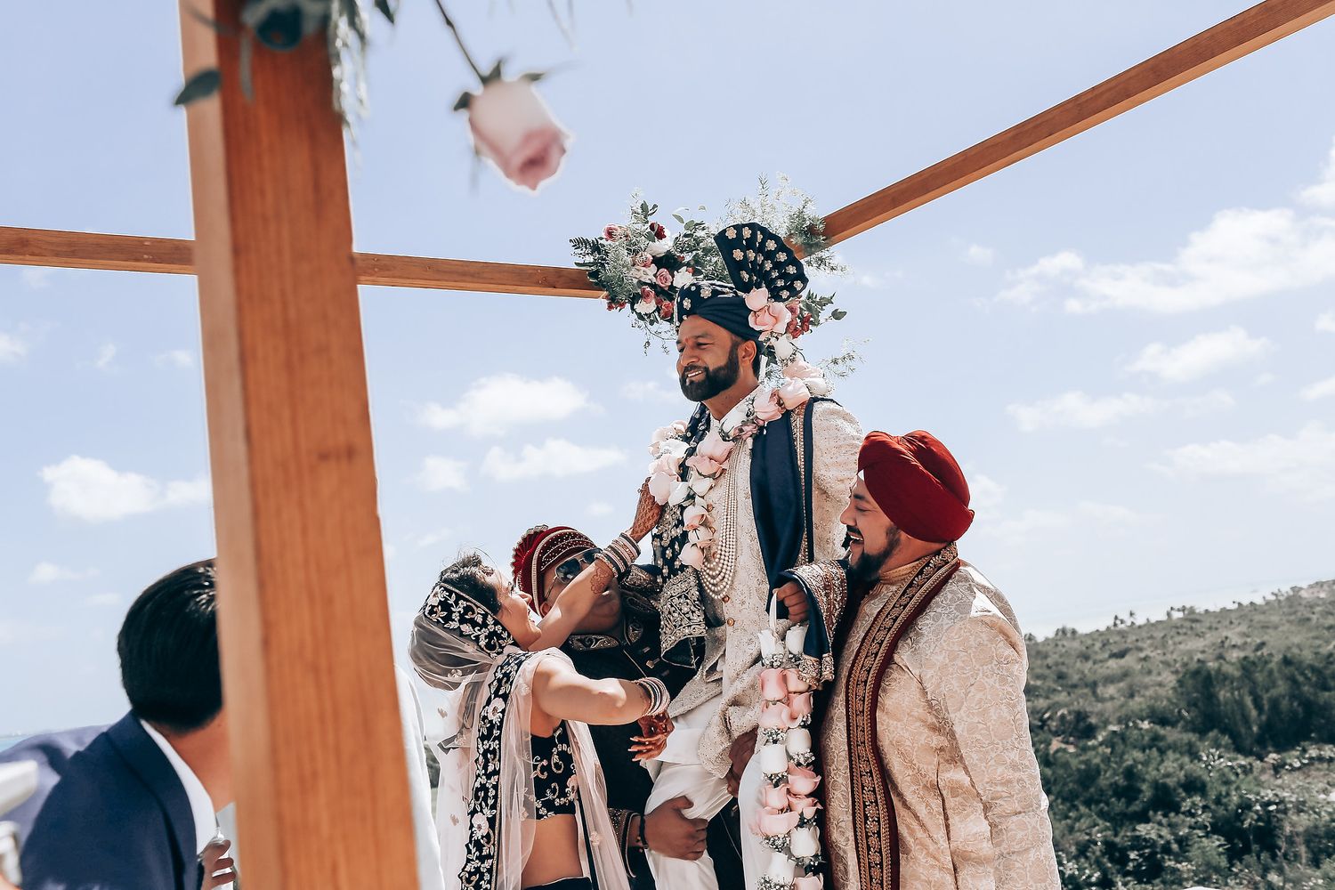 A traditional wedding ceremony takes place under a wooden pergola decorated with white flowers and floral arrangements.