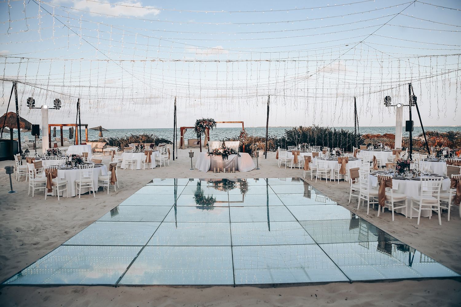 Elegant beach wedding reception setup with white decor and string lights creating a romantic atmosphere at sunset.
