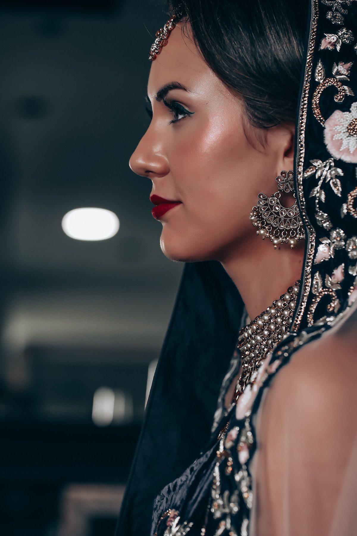 Close-up profile view of a bride wearing ornate traditional Indian wedding jewelry and makeup in dramatic lighting.