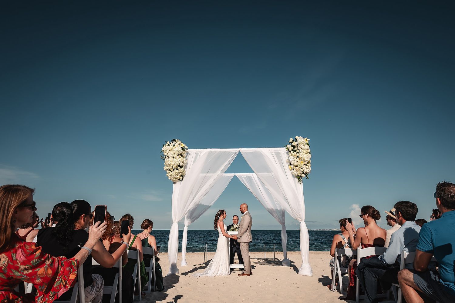 A beach wedding ceremony setup with white fabric arch and chairs against a blue ocean backdrop.