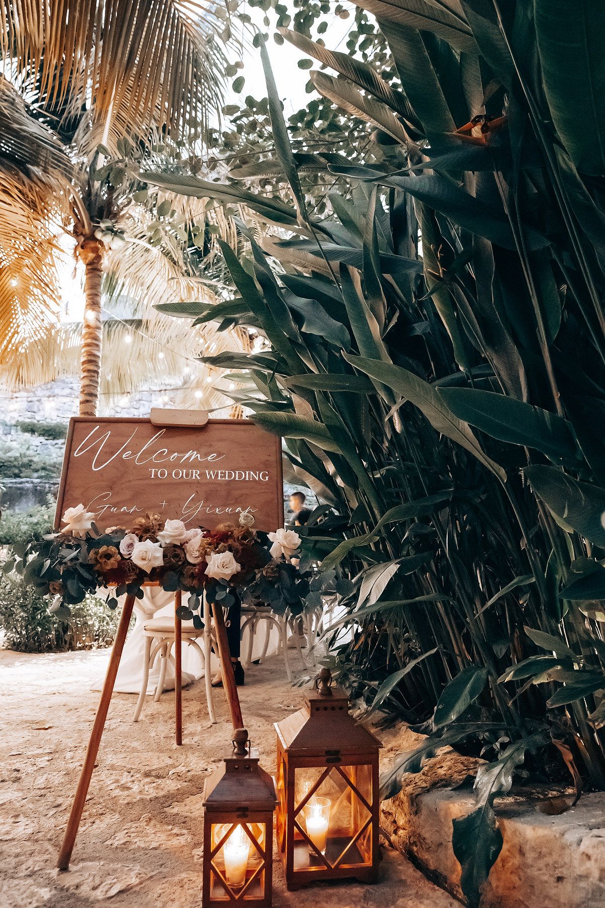 Rustic wooden welcome sign with lanterns and floral decorations at a tropical beach wedding.