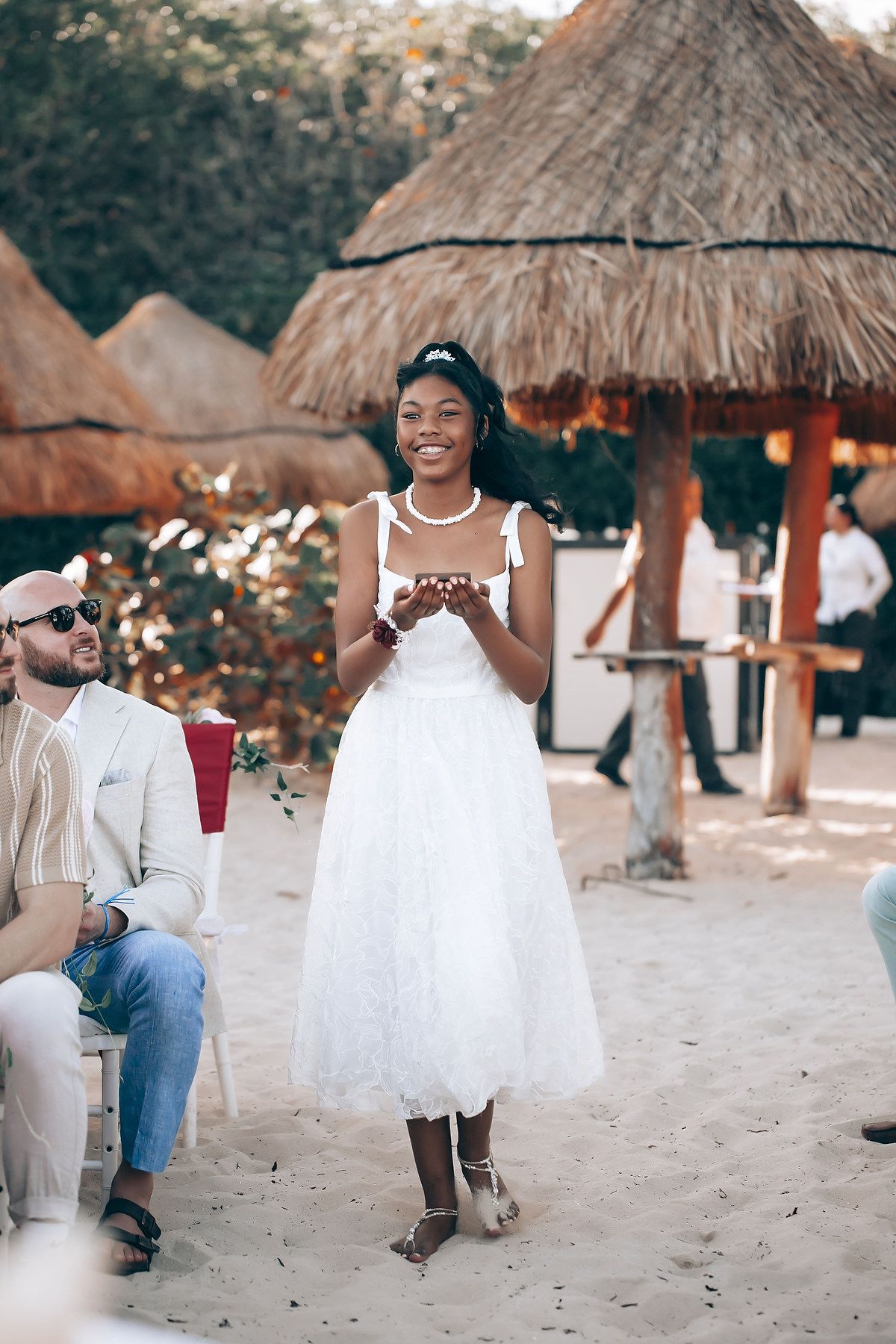 Beach wedding scene with thatched umbrellas and guests in formal attire on sandy shore.