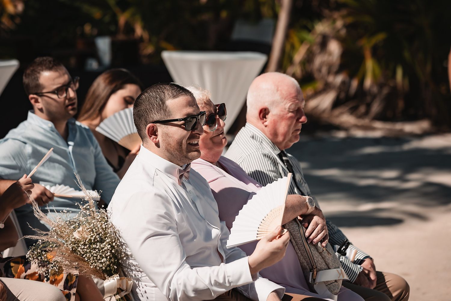 Wedding attendees sit in white chairs during an outdoor ceremony under bright sunlight.