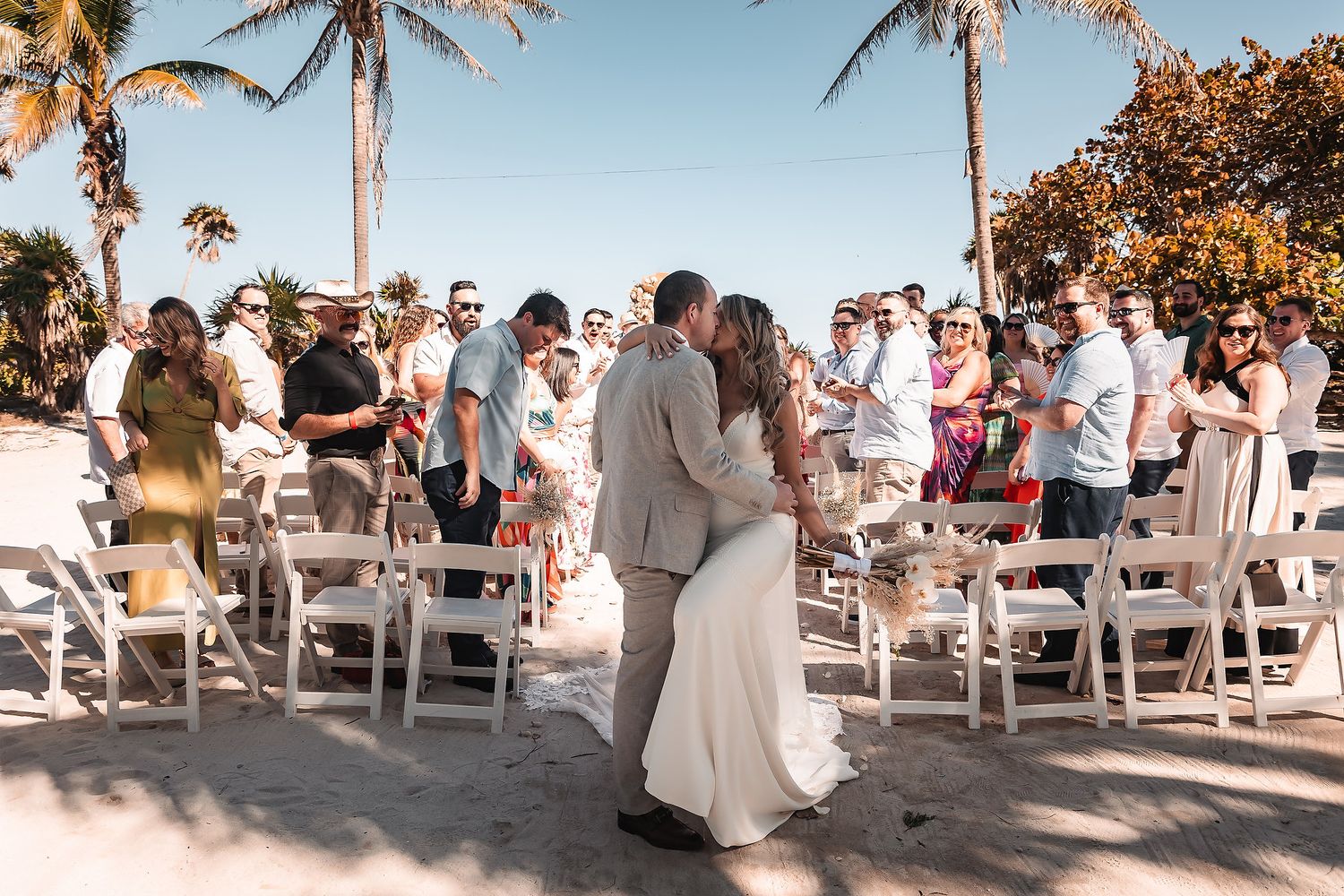Panoramic view of guests seated at a beachside wedding ceremony with palm trees overhead.