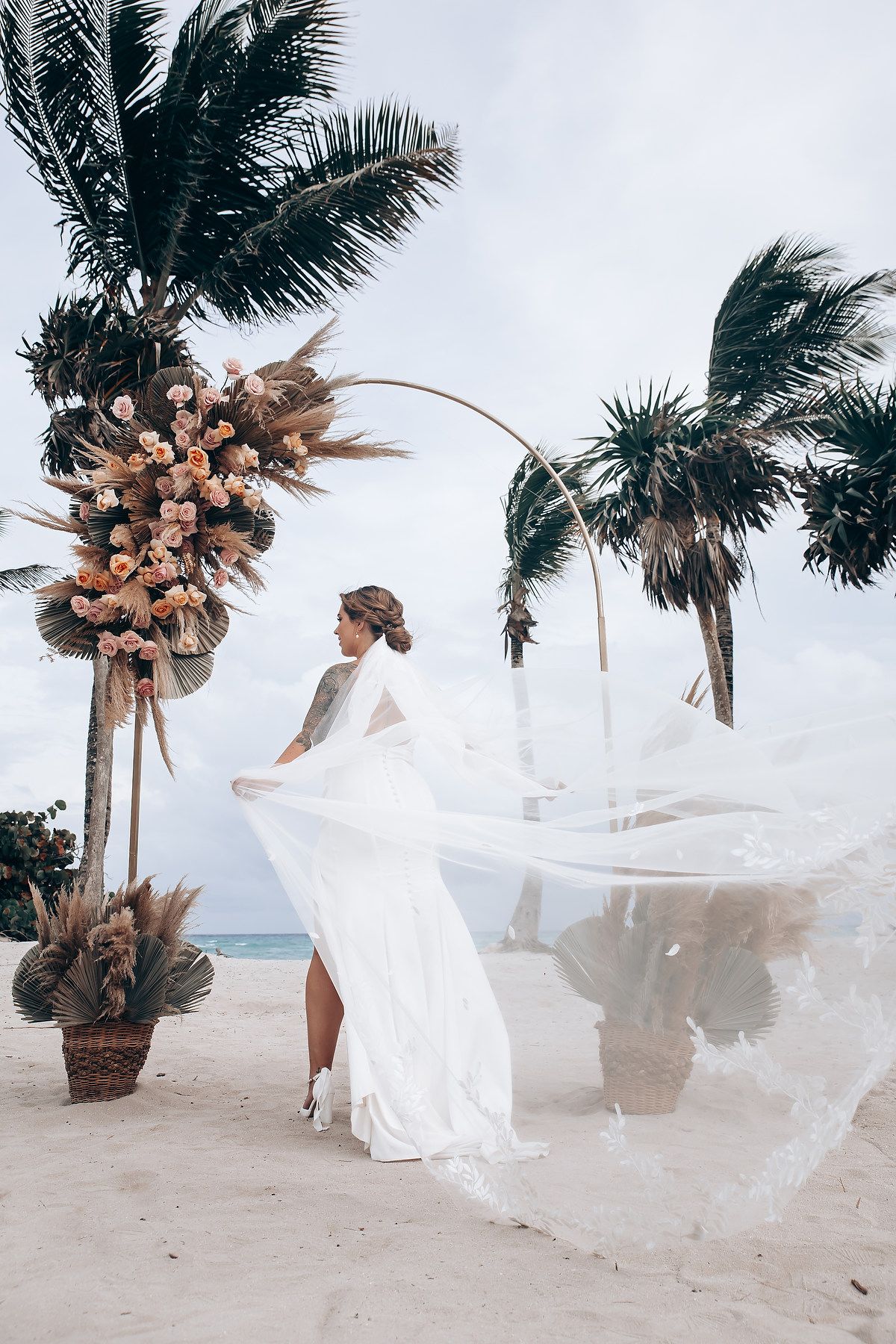 A flowing white wedding dress billows in the ocean breeze against a backdrop of decorated palm trees.
