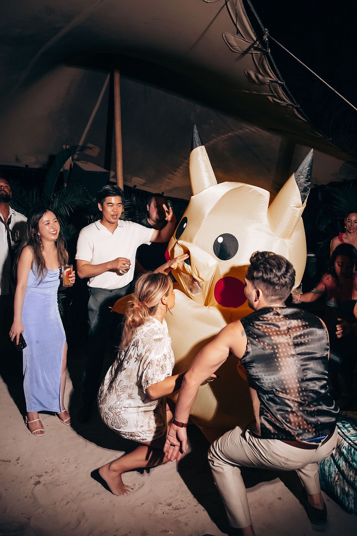 A large Pikachu mascot costume surrounded by party-goers dancing at a nighttime event under a tent.