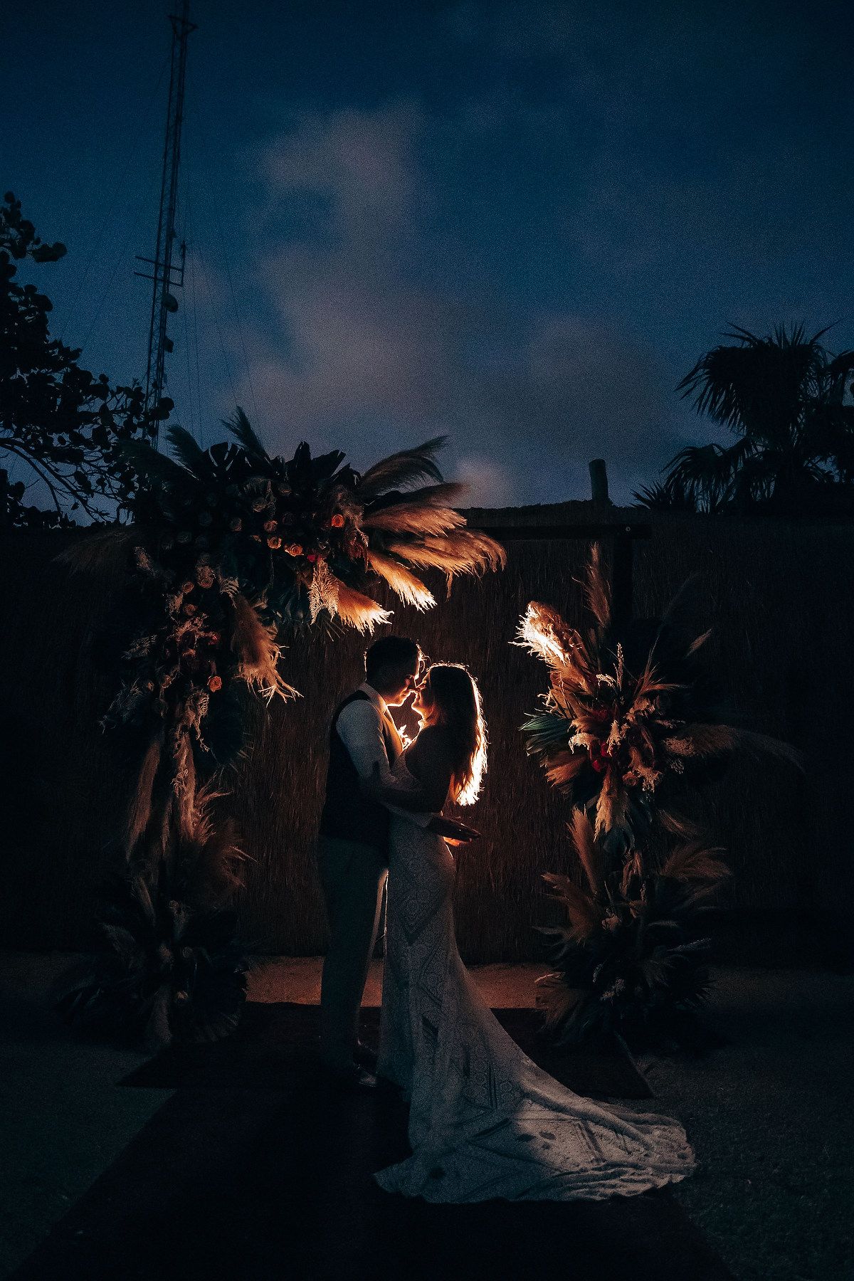 Silhouetted couple embraces under palm trees at twilight with dramatic backlit glow creating romantic ambiance.