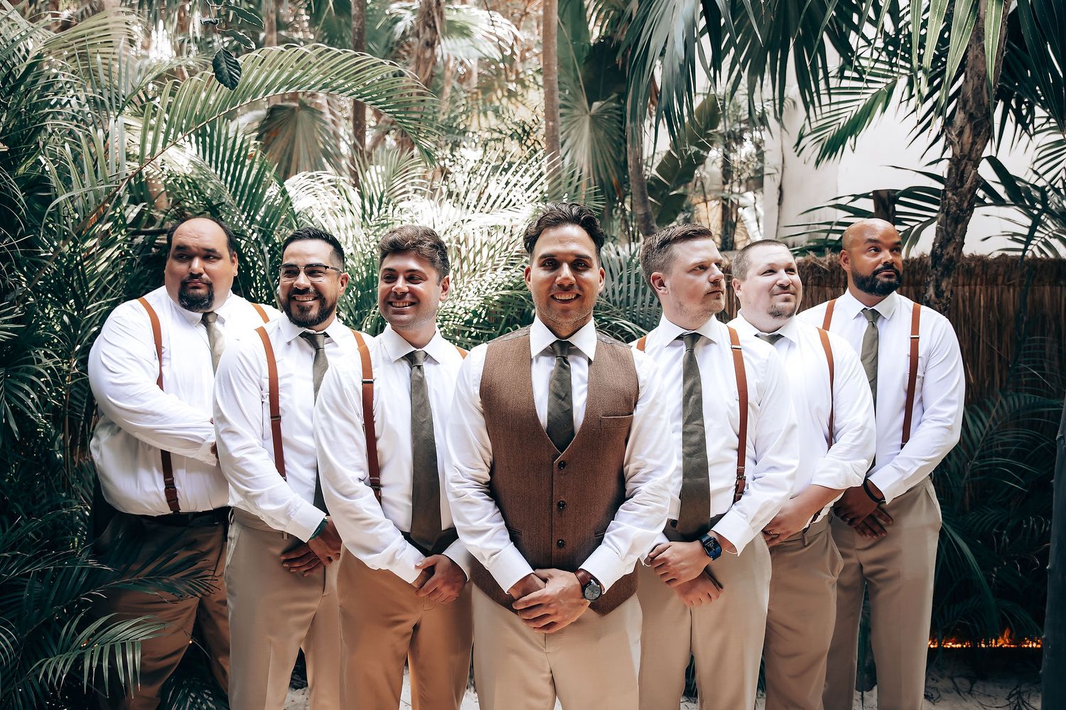 Groomsmen in brown suspenders and white shirts pose together in a tropical outdoor setting.