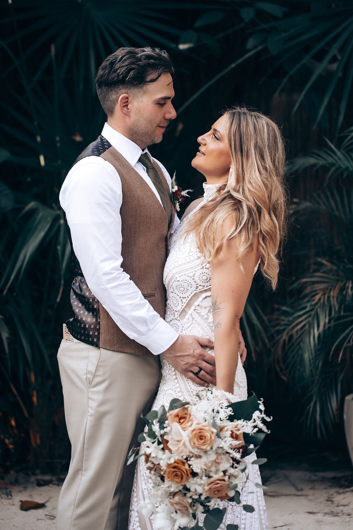 A romantic wedding couple embraces on a beach with tropical foliage and peach-colored bouquet details.