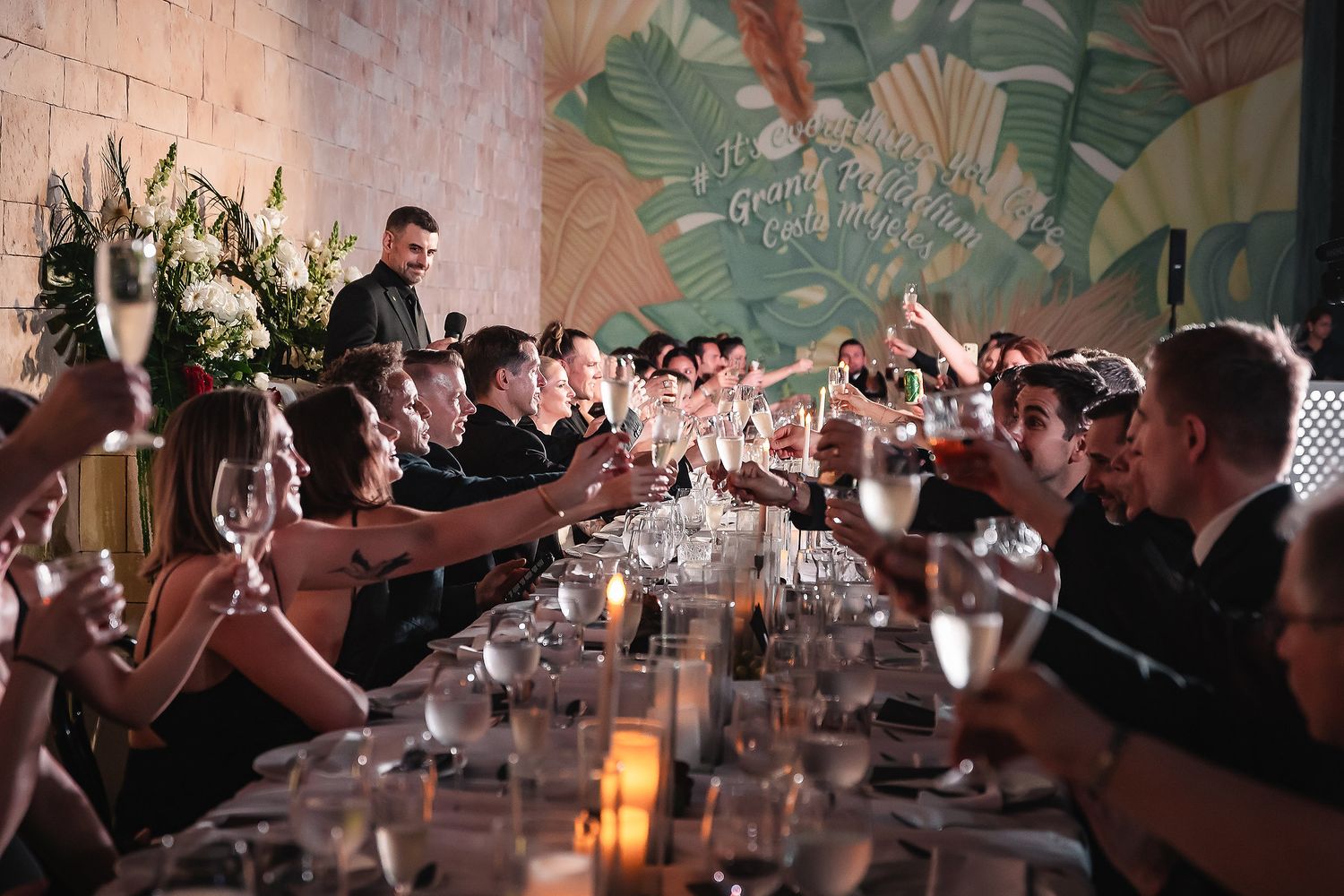 Wedding reception guests raise glasses for a toast at long candlelit table.