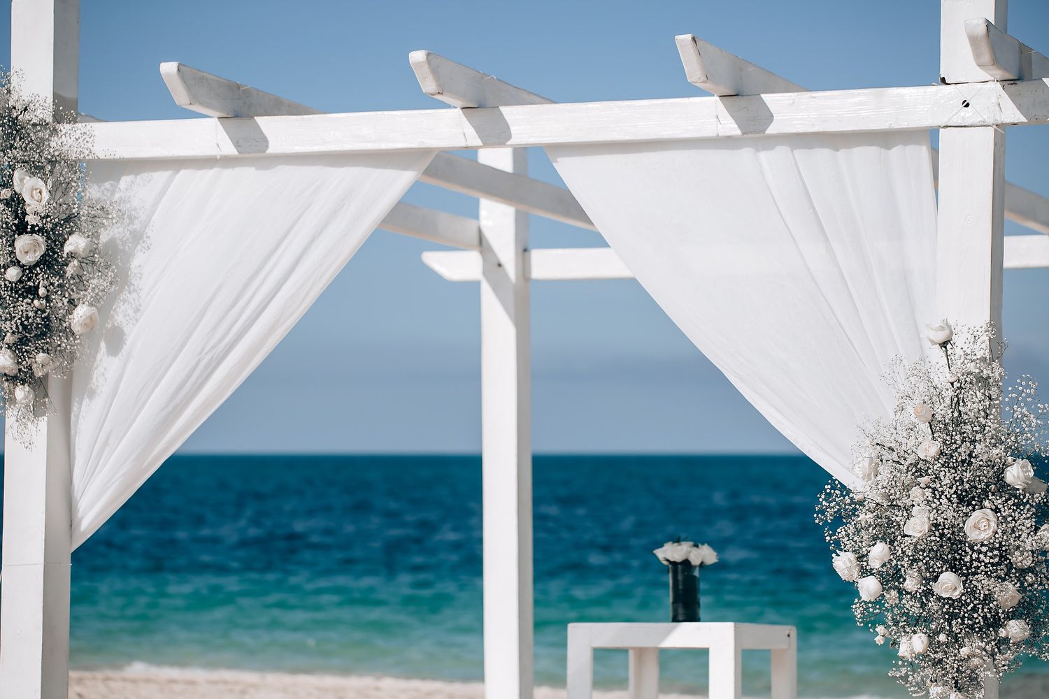 Elegant white wedding arch with flowing fabric and floral arrangements on a pristine beach.