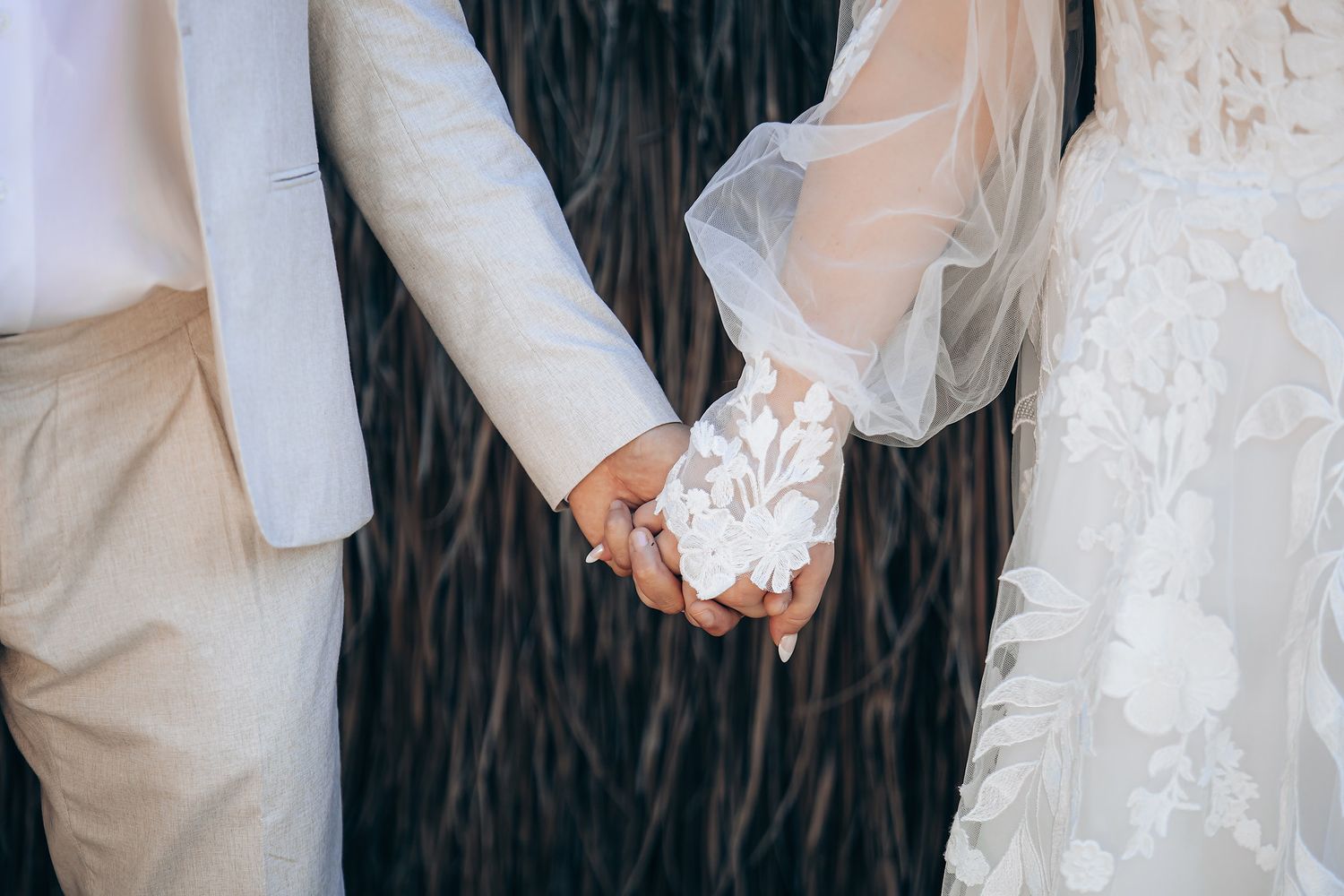 Couple in white wedding attire holding hands against rustic wooden wall backdrop.