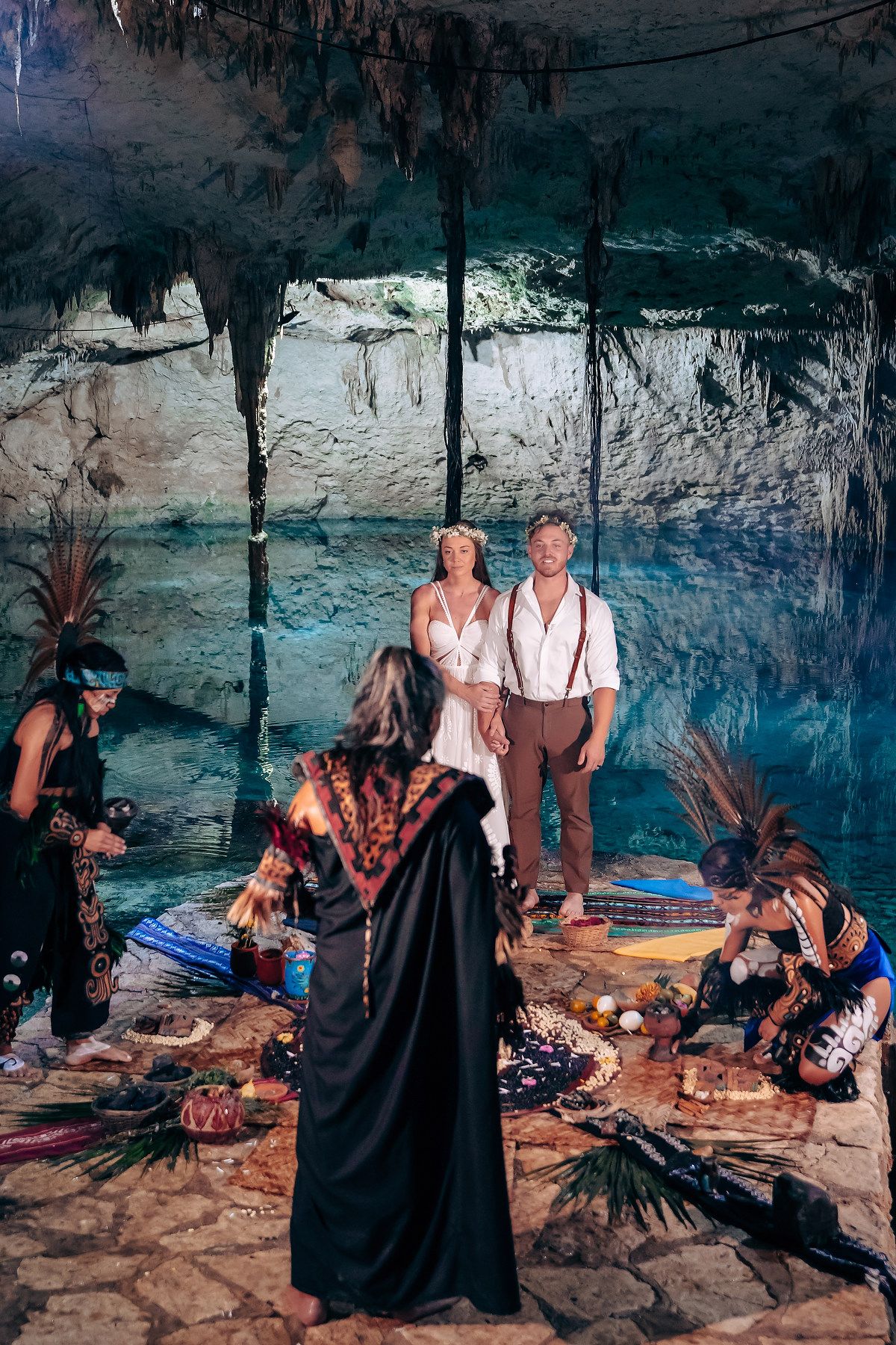 A ceremony takes place in a mystical cenote cave with turquoise water and stalactites hanging from above.