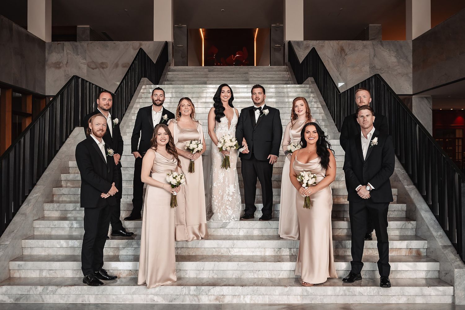 Wedding party in black suits and blush dresses pose elegantly on grand stone steps.
