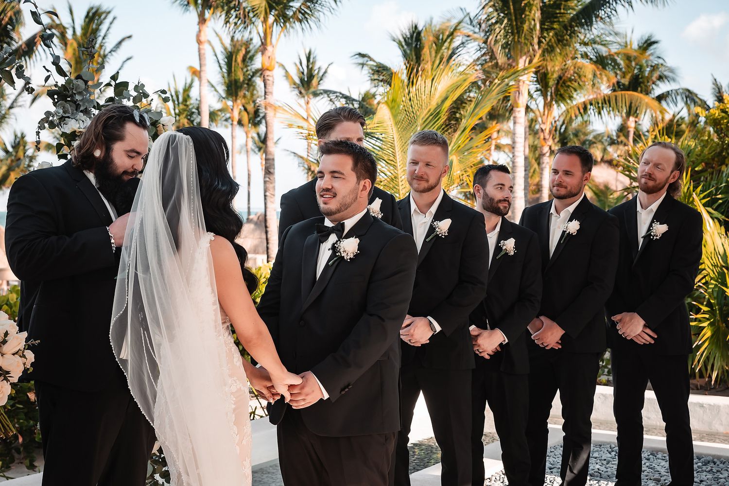 Wedding party laughs together on a sunny day with palm trees and blue sky in the background.