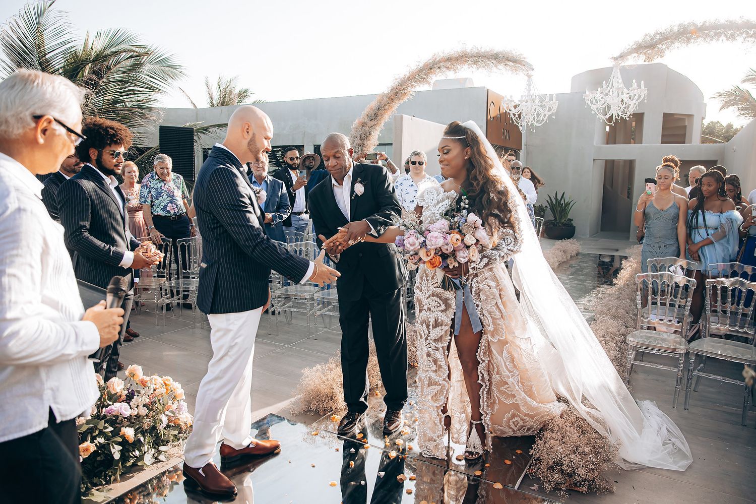 An intimate outdoor wedding ceremony takes place under white arches with palm trees in the background.