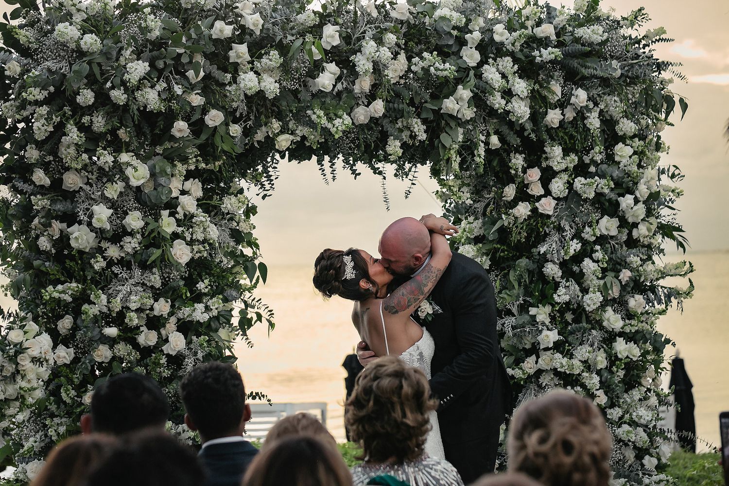 A wedding ceremony takes place under a floral arch at sunset with silhouettes of the couple and guests visible.