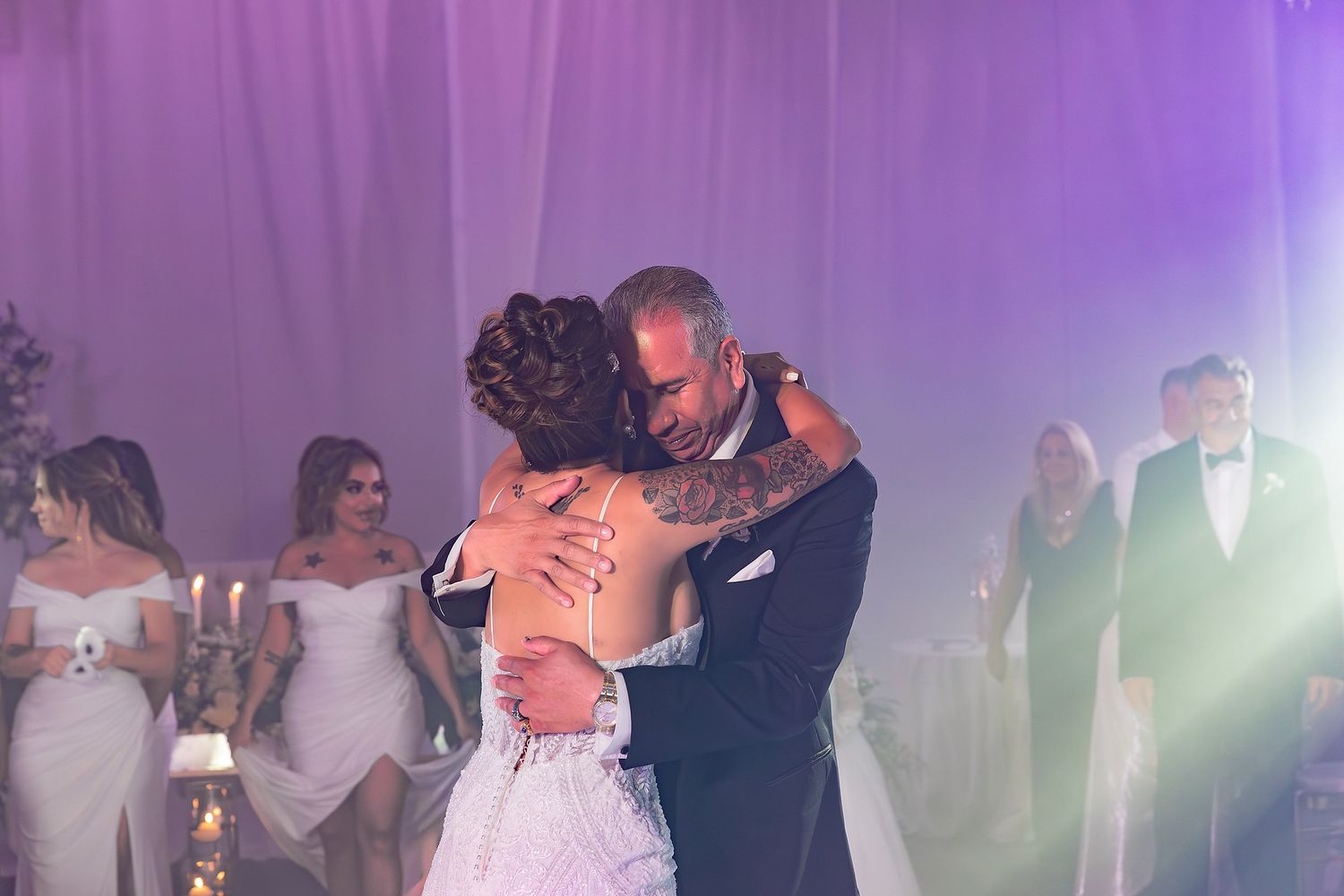 Wedding couple shares first dance on a purple-lit dance floor surrounded by guests at their reception celebration.
