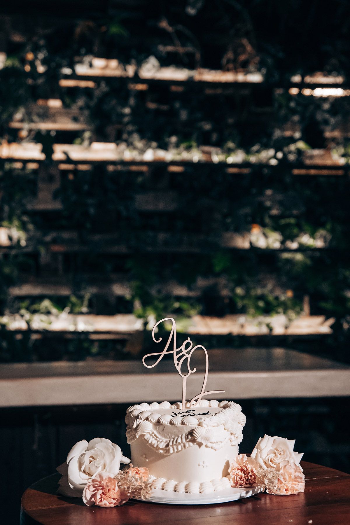 A white wedding cake with decorative topper sits on a dark table with soft lighting and blurred background.