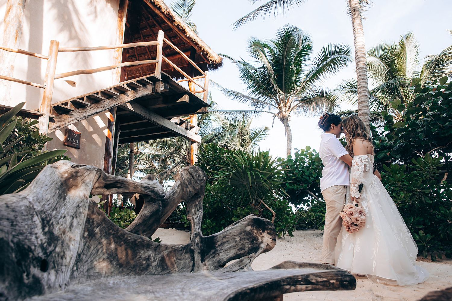 A couple poses near a rustic beach treehouse surrounded by palm trees during a tropical wedding photoshoot.