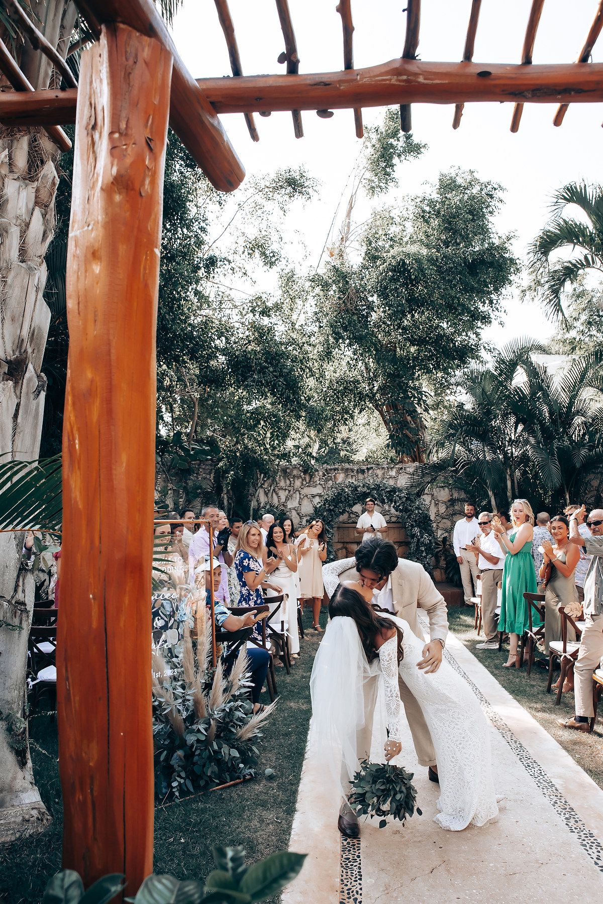 Wedding ceremony under wooden pergola surrounded by tropical landscaping and palm trees.