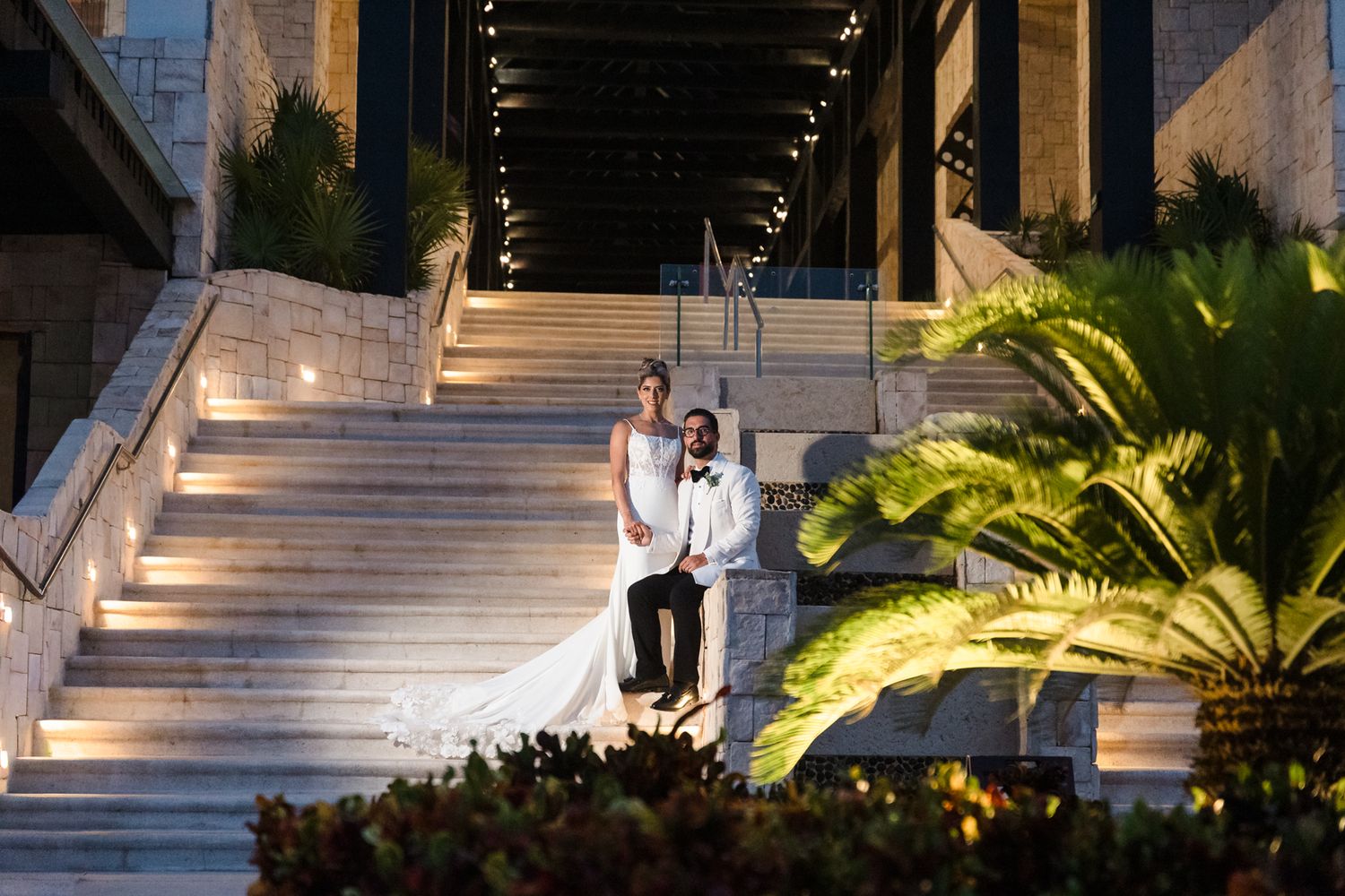 Bride and groom pose elegantly on illuminated stone steps surrounded by tropical greenery at night.