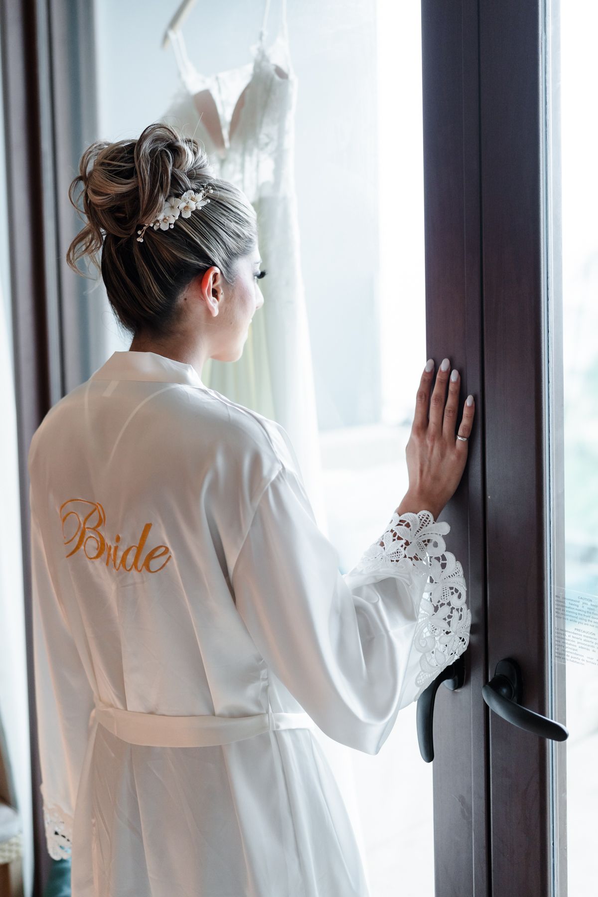Bride in white satin robe with gold script looks at wedding dress hanging by window.
