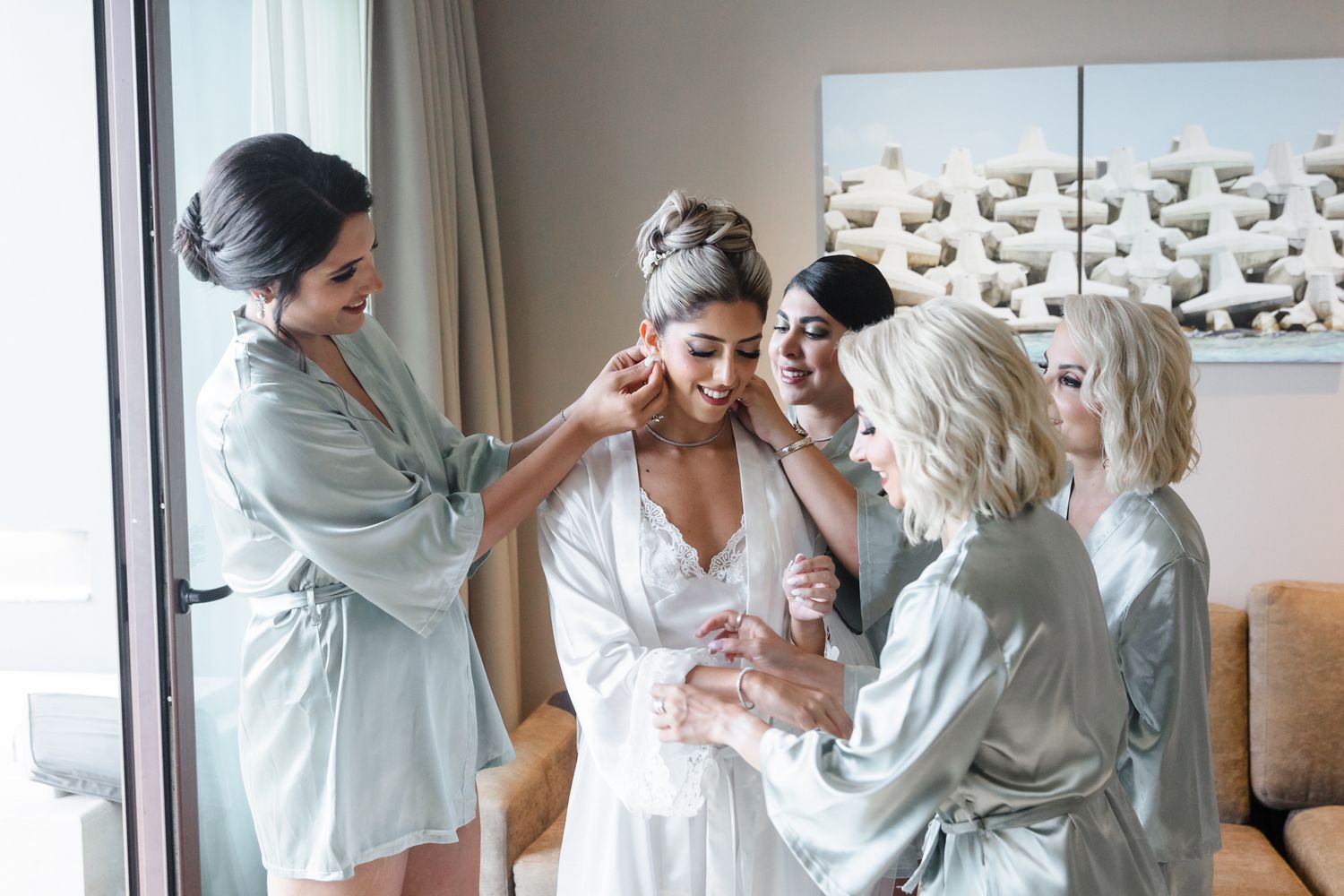 Bridesmaids in sage robes help bride with earrings and dress on wedding morning.