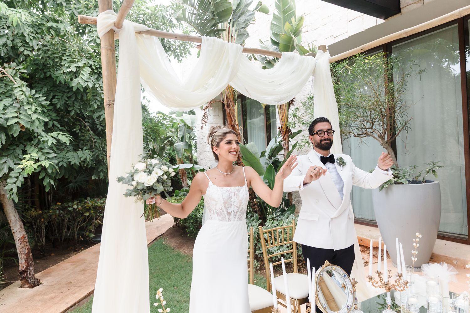 Bride and groom celebrating under a white draped wedding arch surrounded by lush greenery at an outdoor ceremony.