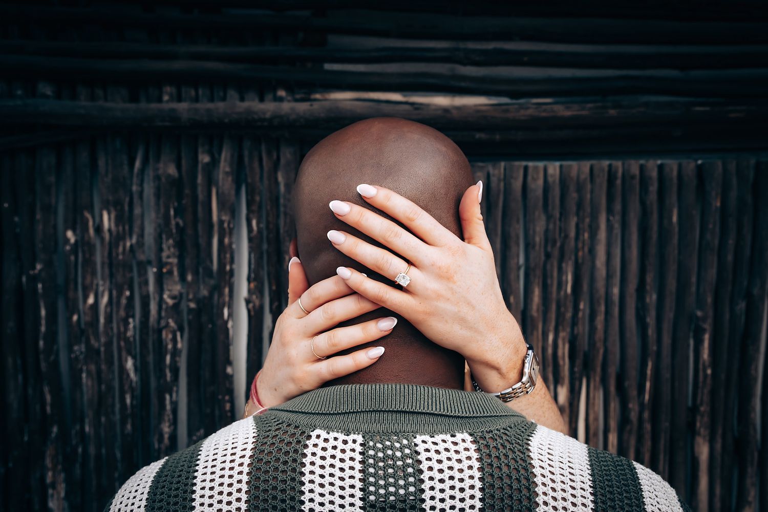 Woman placing her hand on a bald mans head from behind near a wooden fence.